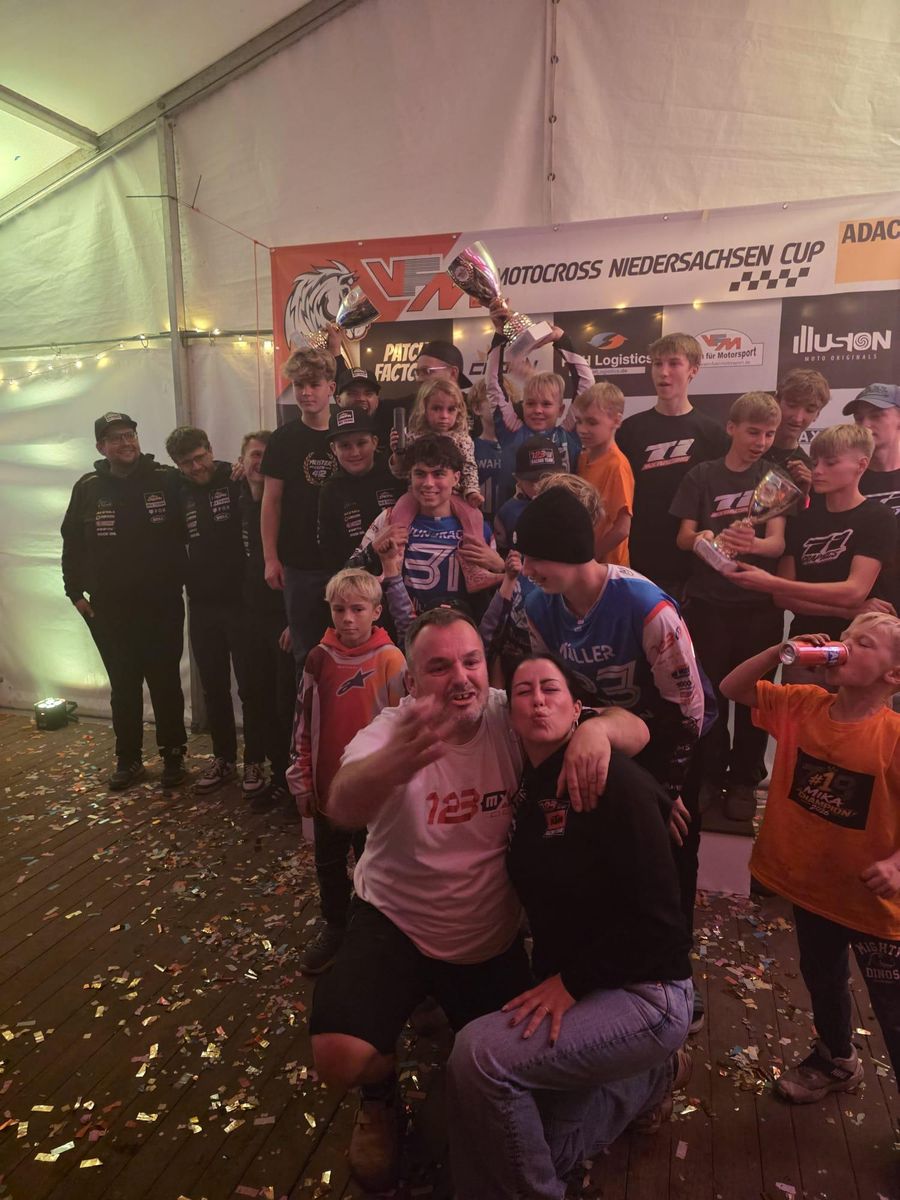Fahrer, Eltern und Betreuer feiern im Festzelt vor dem Banner des VFM Motocross Niedersachsen Cup mit Pokalen und Konfetti