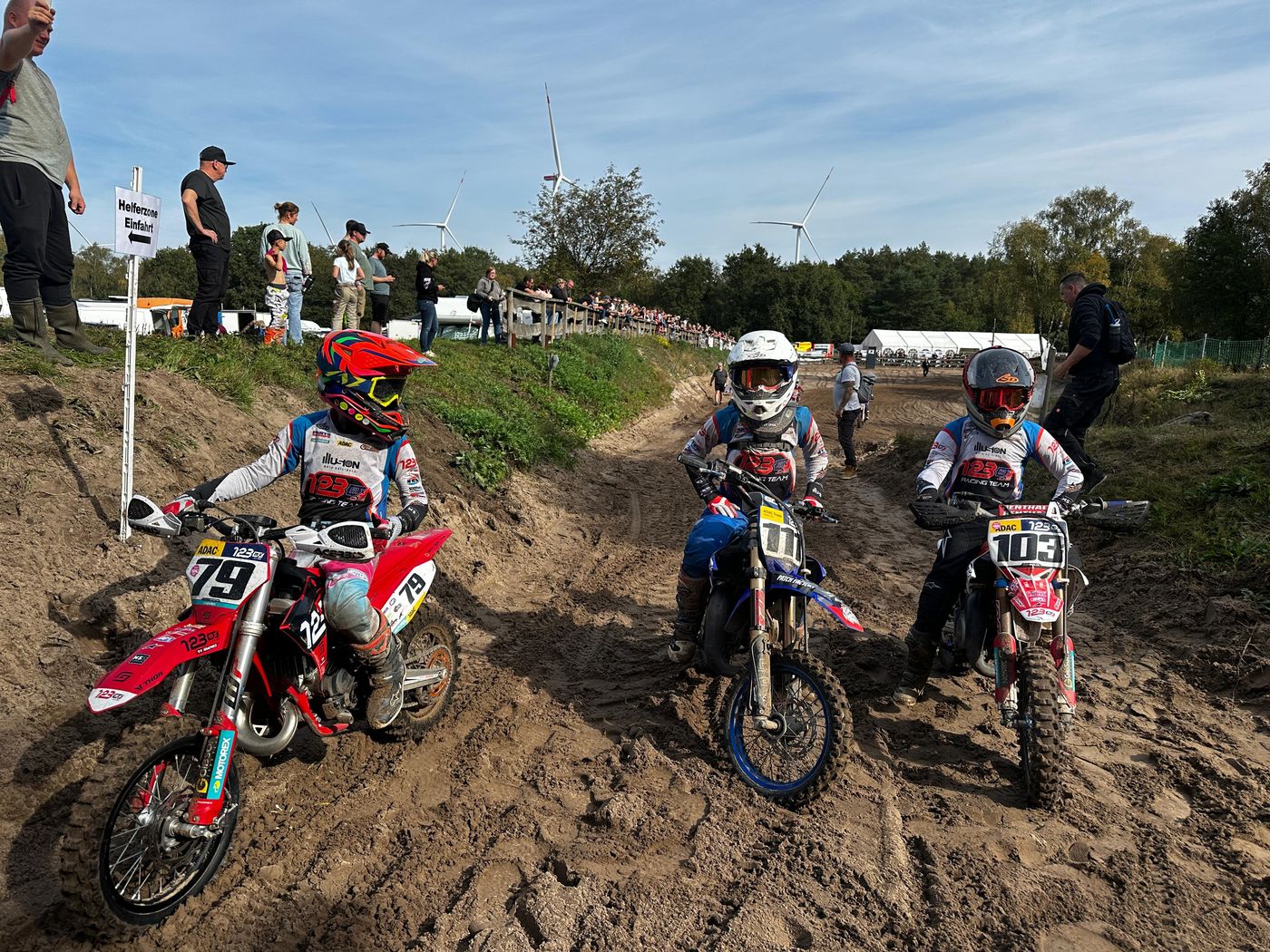 Drei junge Motocrossfahrer mit den Startnummern 79, 12 und 103 warten auf ihren Motorrädern im Sand vor dem Start, im Hintergrund Zuschauer und Windräder