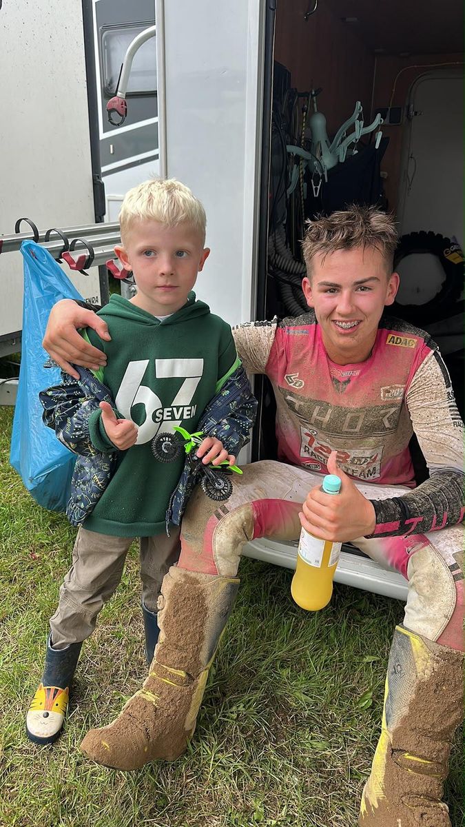 Mio Müller im verschmutzten 123MX-Racing-Team-Trikot sitzt am Wohnmobil und legt den Arm um einen kleinen Jungen, der ein Spielzeug-Motorrad hält