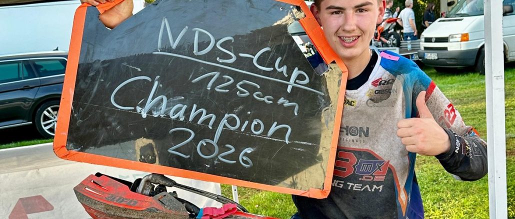 Mio Müller hält eine Tafel mit der Aufschrift NDS-Cup 125ccm Champion 2026 und zeigt den Daumen nach oben, vor ihm sein Motorrad mit der Startnummer 1