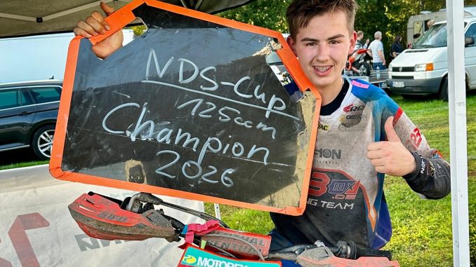 Mio Müller hält eine Tafel mit der Aufschrift NDS-Cup 125ccm Champion 2026 und zeigt den Daumen nach oben, vor ihm sein Motorrad mit der Startnummer 1