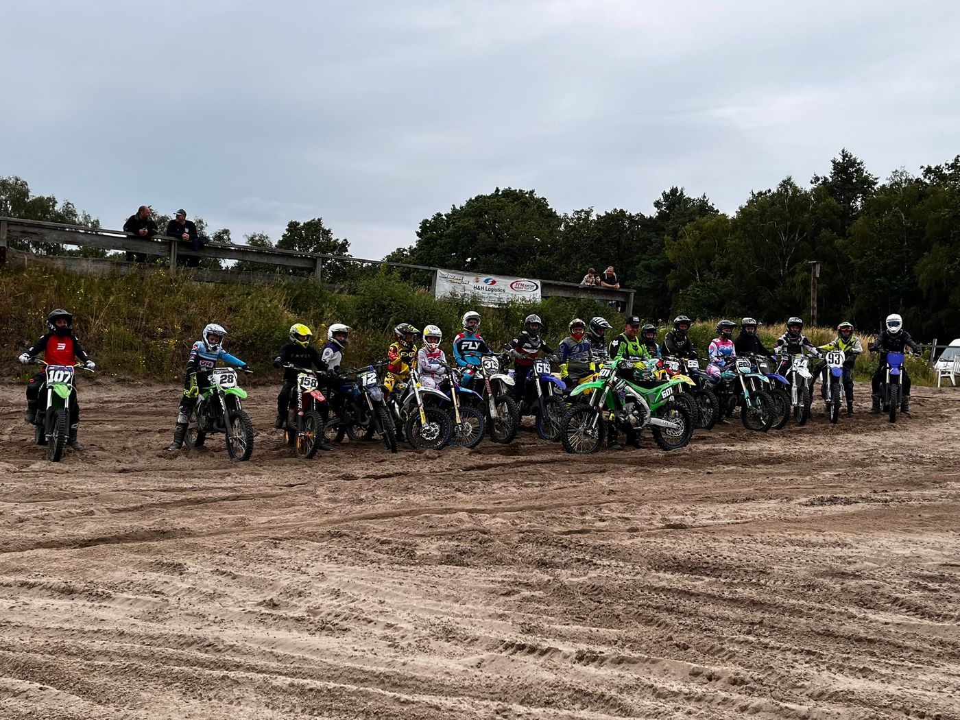 Rund zwanzig Fahrerinnen und Fahrer stehen mit ihren Motocross-Motorrädern nebeneinander im tiefen Sand