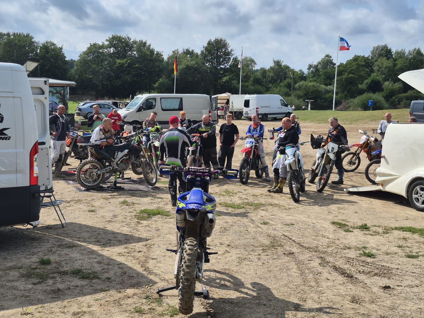 Erwachsene Trainingsteilnehmer stehen mit ihren Motorrädern im Halbkreis und hören bei einer Besprechung im Fahrerlager zu